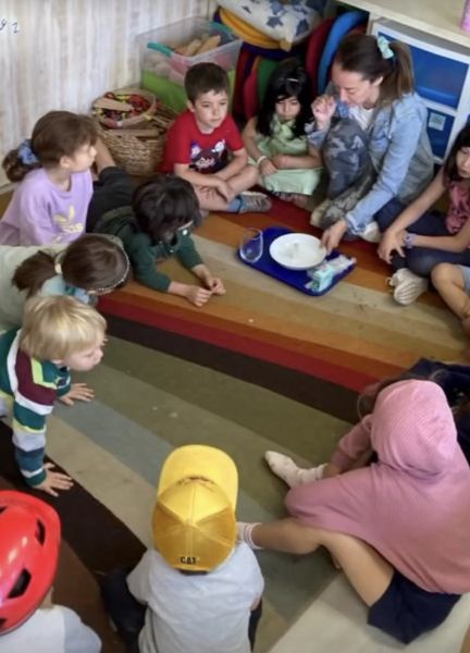 Una adulta acompaña un experimento en grupo con niños sentados en una alfombra, Sommerzeit 2024